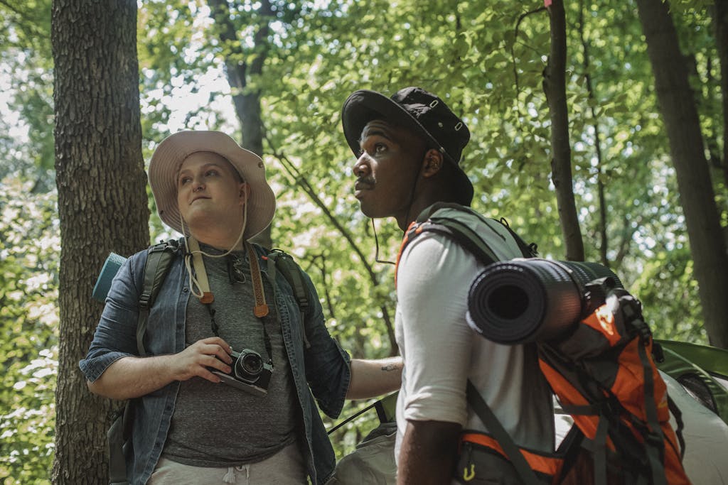 Plus size guy with photo camera and black young bearded man with backpack and tourist mat standing near tent and looking up